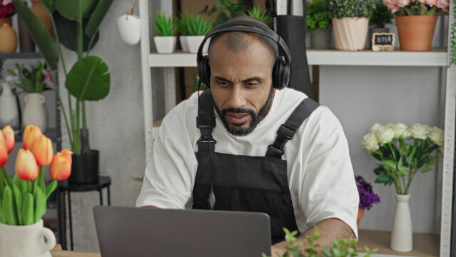 Man wearing apron and headset typing on laptop with smiling face in flower shop; cheerful online service.