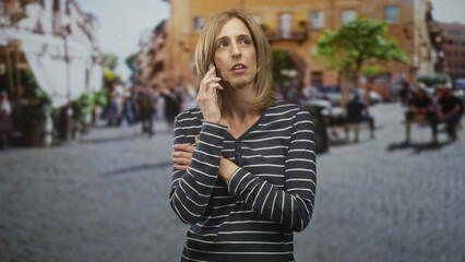 Fototapeta premium Woman holding smartphone to ear with arms crossed in a cobblestone street plaza, eyes closed and pensive expression; concern waiting.
