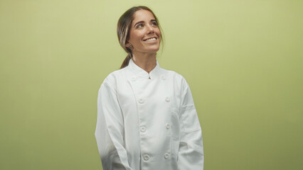 Fototapeta premium Young hispanic woman chef smiling with face visible and hands clasped, wearing white chef coat in studio against green wall; confidence culinary craft.