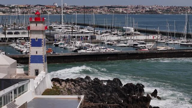 Aerial view of Farol Museu de Santa Marta with its blue and white stripes overlooking the marina and crashing waves on the rocky shore in Cascais, Lisboa, Portugal.