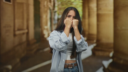 Fototapeta premium Woman with fists raised covering her face in a building colonnade walkway, hands tense and protective; anxiety resilience guarded determination.