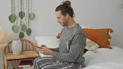 Fototapeta premium Man sitting on bed in gray loungewear reaching with right hand for red smartphone on wicker nightstand beside pillows and macrame wall hanging in studio; calm morning.