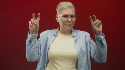 Naklejka premium Woman making air quotes with both hands, wearing glasses and a light button shirt in a red studio; playful skepticism.