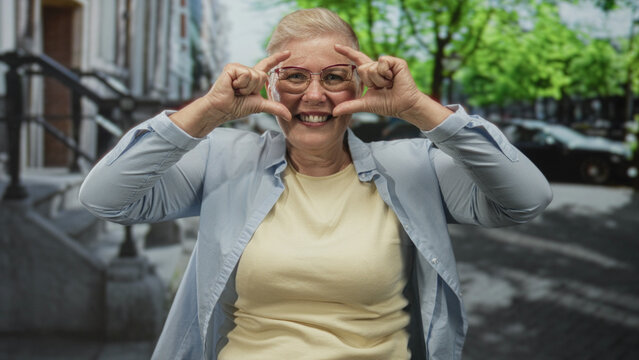 Middle aged woman smiling and framing her face with both hands on a busy street by stoop steps and parked cars; playful joy.