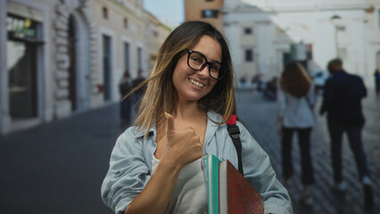 Fototapeta premium Young woman holding books and flashing a peace sign with her hand on a cobblestone street near a plaza and buildings; playful confidence.