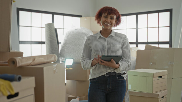Woman tapping tablet amid packed moving boxes, bubblewrap and an ironingboard in a building, checking inventory and scheduling tasks; focused planning.