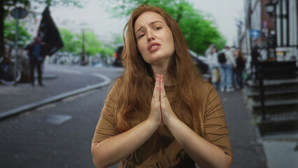 Fototapeta premium Woman in brown dress with hands pressed in prayer gesture on city street amid blurred pedestrians; prayer hope.