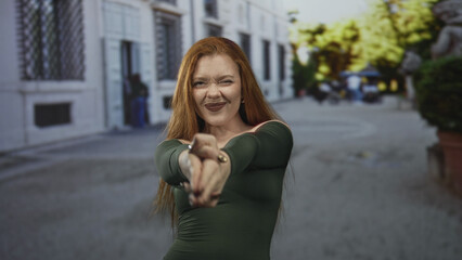 Obraz premium Redhead woman with bare shoulder making finger gun gesture on a cobbled street; playful confidence.