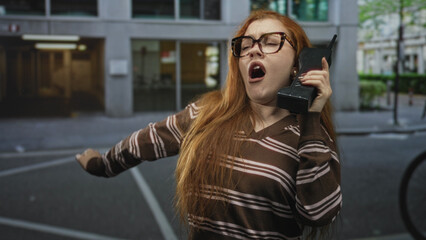 Obraz premium Young redhead woman singing into a retro cellphone on a city street, arm outstretched and mouth open, wearing brown striped sweater; playful nostalgia.