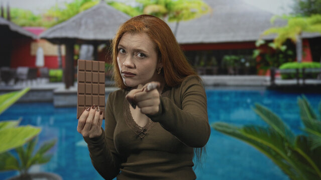 Woman redhead holding large chocolate bar pointing index finger at camera in studio pool villa set; accusation temptation warning.