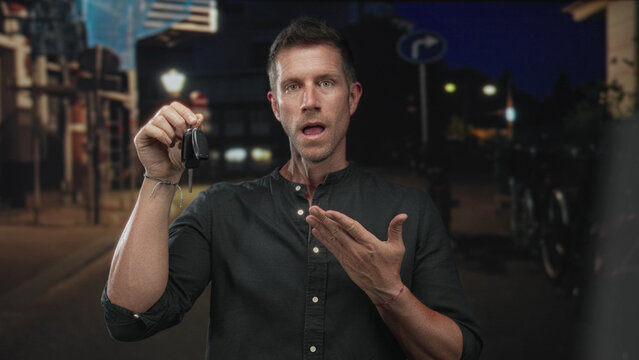 Man young caucasian holds car key fob with raised hand and points to keys on street at night with open mouth; surprise lost item.