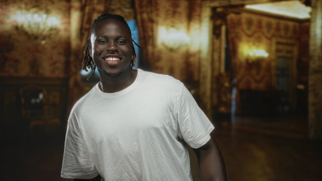 Man smiling, wearing a white t shirt with earring and visible dreadlocks, standing inside an ornate museum building with warm lighting; joyful confidence.