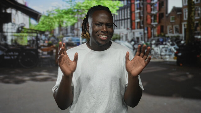 Man with hands raised, palms out on a street, eyes squinted and wincing while wearing a white t shirt; awkward hesitation.