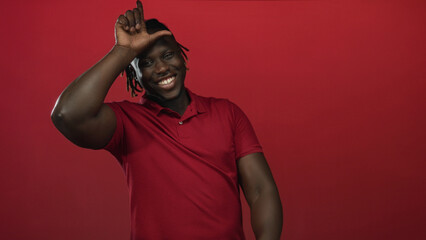 Obraz premium Man with hand on forehead forming l gesture and pointing finger toward camera while smiling in a red studio backdrop and wearing a red polo shirt; playful confidence.