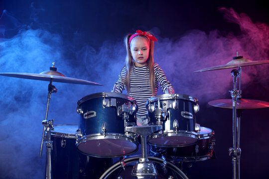 Little girl in bandana learning how to play drums in color lights and smoke against dark background