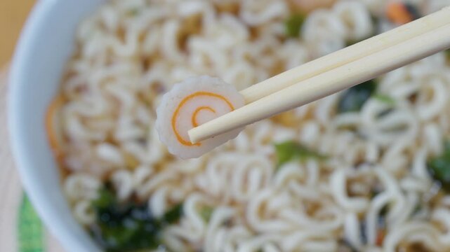 Narutomaki fish cake held with chopsticks over instant ramen noodles close up asian food detail quick meal concept japanese cuisine takeaway style. 