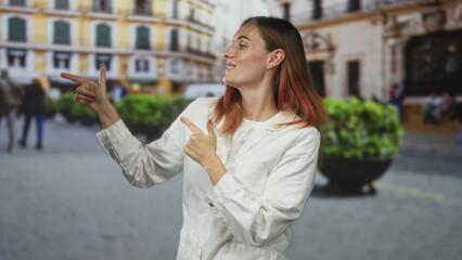 Fototapeta premium Young redhead woman in white coat gestures finger guns with both hands, smirking and pointing across a city street framed by historic buildings; playful confidence.