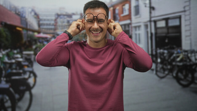 Young hispanic man with moustache and round glasses tugs his ears with fingers, smiling in a city street with parked bicycles and storefronts; playful joy.