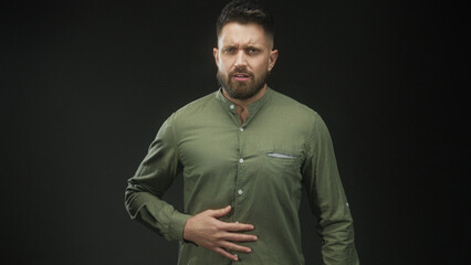 Obraz premium Man, bearded and young, clutching stomach with both hands, wincing and leaning forward, wearing green button shirt in studio; pain vulnerability.