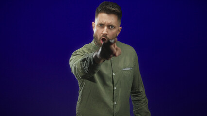 Obraz premium Hispanic man with beard thrusts clenched fist toward camera in studio with olive shirt, stern gaze and scowl; anger confrontation.