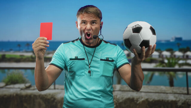 Referee man holds red card high and cradles soccer ball on a street with headset and whistle, stern expression and outstretched arms; authority fairness.