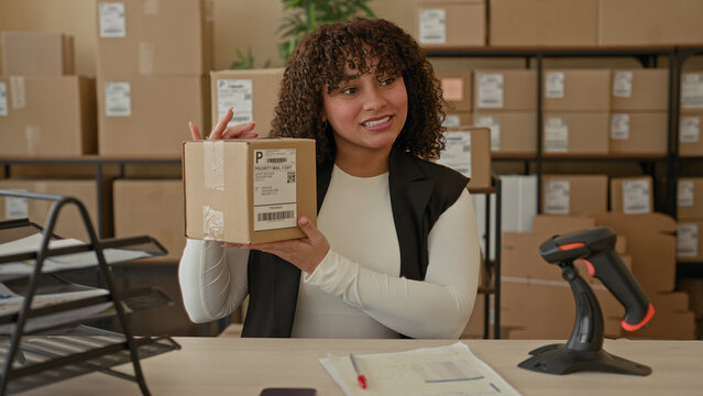 Woman holding package and gesturing with hand in a building delivery office amid stacked cardboard boxes and scanner; confidence entrepreneurship success.