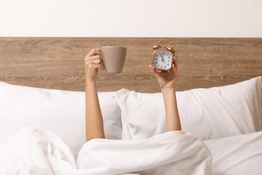 Female hands with alarm clock and cup of coffee in bed at home