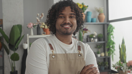 Fototapeta premium Man florist wearing apron holding flowers, arms crossed and smiling near potted plants and vases by a bright window in a shop building; pride craft.