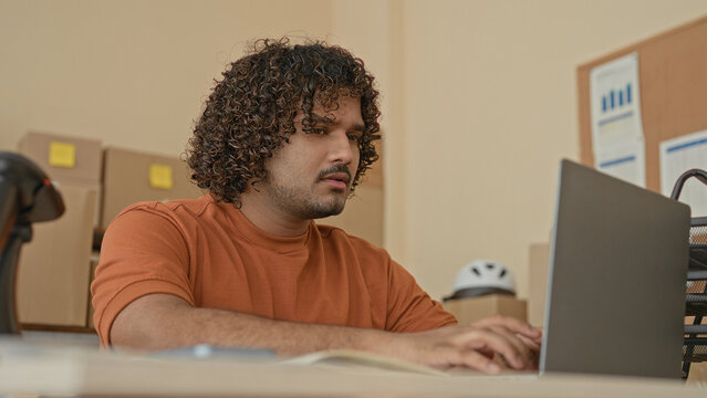 Man thinking with hand on chin at laptop among cardboard boxes and parcels, reviewing orders at a small business desk in building; planning.