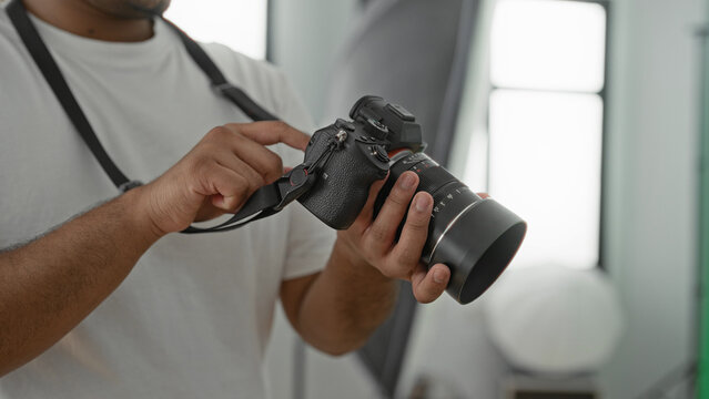 Young indian photographer adjusts camera dial with hands in studio near softbox and window; concentration precision craft.