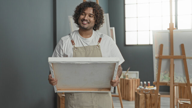 Man holding blank canvas with both hands, smiling and cradling it, wearing apron, in art studio with easels, paint bottles and wooden stools by window; creative joy.