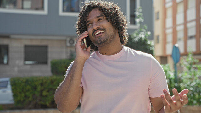 Man holding smartphone to ear, smiling and gesturing on a sunny street in front of buildings; friendly conversation cheerful.