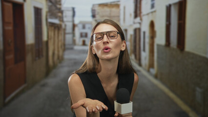 Obraz premium Woman reporter holding microphone wearing glasses reaching forward with open hand while blowing a kiss on an old town cobbled street; playful charm.