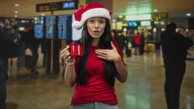 Young woman in santa hat smiling softly holds a red mug with one hand and places the other hand on her chest at airport; holiday warmth giving.