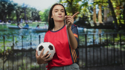 Naklejka premium Young woman holding soccer ball, pointing index finger up, wearing red spain jersey and jeans in studio with park backdrop; pride support.