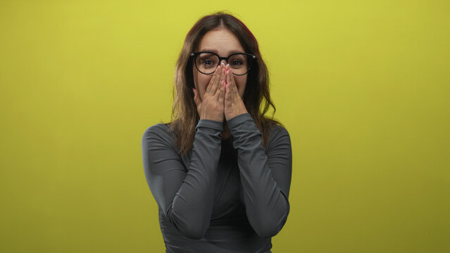 Hispanic woman wearing glasses covering mouth in lime green studio while smiling and laughing; surprise candid moment.