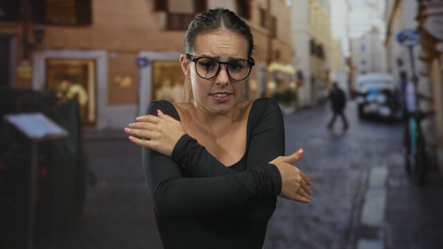 Woman rubbing hands and squinting with glasses on a narrow city street lined with old buildings; unease.