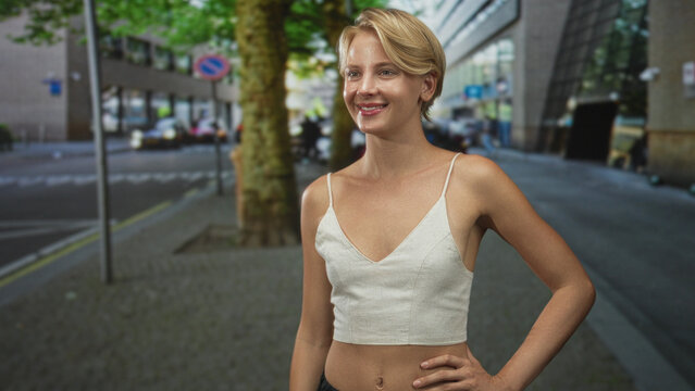 Blond woman extends arm palm up on a city street, hand offered as if to shake or receive, left hand on hip, smiling toward unseen person; friendly invitation.