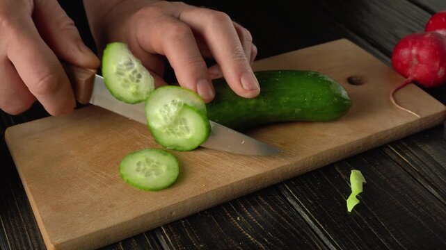 Slicing fresh cucumber into thin rounds on a wooden cutting board with radishes and green leaves visible in the background