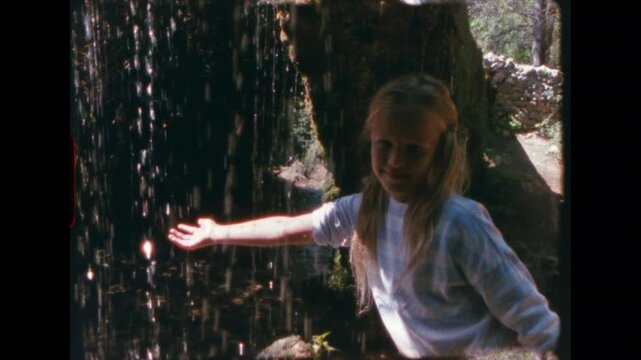 Young girl leaning forward and extending arm into falling water stream beside ancient stone wall, magical childhood moment captured on super 8 vintage film with warm grain