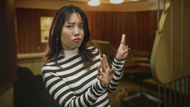 Woman with palms outward hand gesture and pursed lips in a building booth, wearing striped sweater and visible ring as she pushes away with fingers; disapproval dismissal.