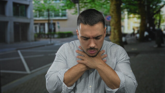 Young hispanic man with furrowed brow and closed eyes clutching throat on urban street corner; distress.