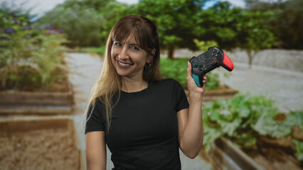 Woman holds wireless game controller near shoulder in park with green trees and raised planter beds, smiling at camera  play fun. © Krakenimages.com