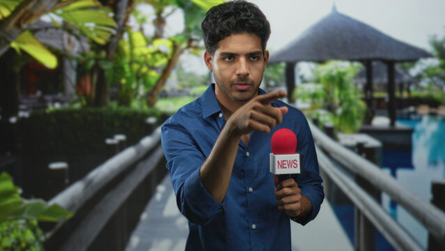 Man holding red news microphone and pointing finger toward camera on a resort walkway in building; live reporting confidence.
