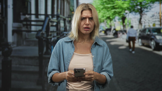 Woman holding smartphone with hands, typing and frowning while looking at the screen on a street in the city; concern.
