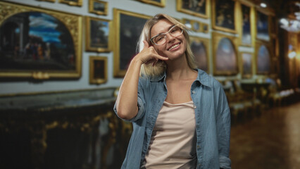 Fototapeta premium Woman makes call hand gesture to her ear while smiling in a museum building filled with framed paintings; playful curiosity.