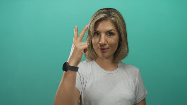 Young blonde woman displays victory sign with raised hand in turquoise studio; triumph confidence spirit.