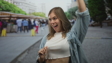 Fototapeta premium Blonde woman dancing with exposed stomach and raised arms in busy street market by souvenir stalls; happiness.