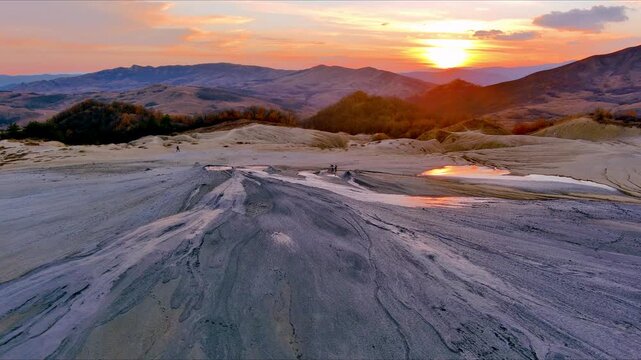 Aerial view of Vulcanii Noroiosi Paclele Mici mud volcanoes with grey cracked earth and rolling hills under a golden sunset sky in Scortoasa, Buzau County, Romania.