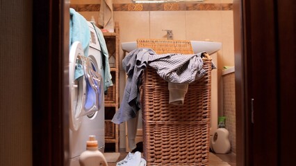 Bathroom interior showing an overflowing wicker laundry basket, clothes in a washing machine, and cleaning supplies, representing household chores and general mess © Aliaksandr